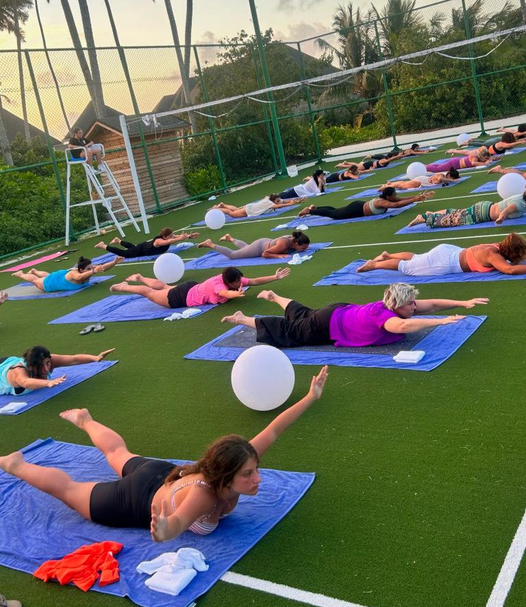 Group of people doing yoga outdoors with palm trees and a sports field in the background.